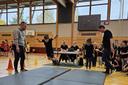 Kind springt in einer Turnhalle mit geschlossenen Beinen nach vorn, darunter liegen weiche Matten. Es ist ein Wettkampf. Weitere Personen schauen zu.